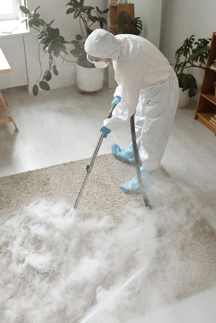 A professional cleaner wearing a grey jumpsuit is using a yellow and black industrial vacuum cleaner on a dark carpeted floor in an auditorium or conference room. The room features tiered seating with