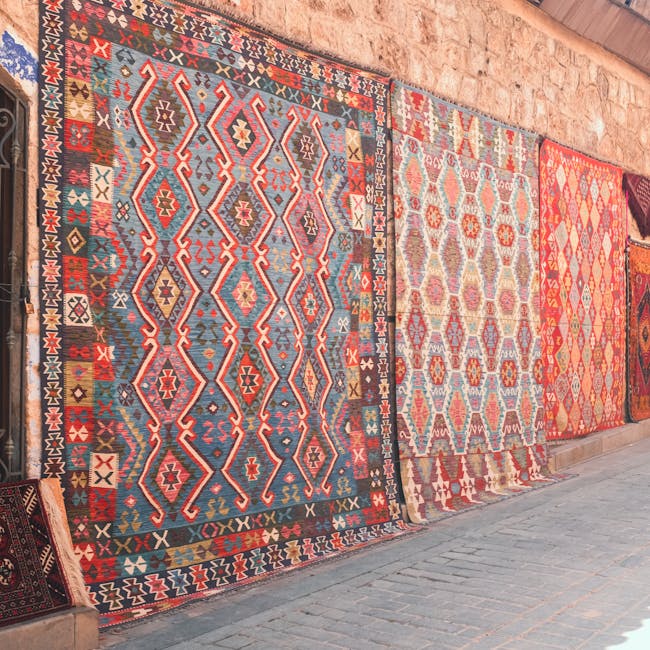 A row of traditional patterned rugs hanging vertically on a brick wall exterior, showcasing intricate geometric designs in vibrant colors such as red, blue, beige, and orange. The rugs vary in size an