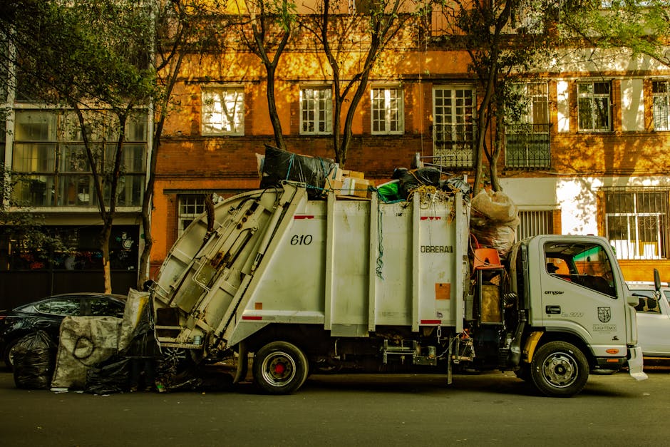 A row of black wheelie bins with orange and blue lids, situated against a red brick wall on a concrete surface, representing waste disposal guidelines in Hackney. The bins are clean and neatly aligned